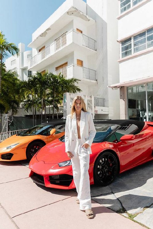 Woman in white blazer standing confidently between three luxury sports cars in Art Deco Miami Beach